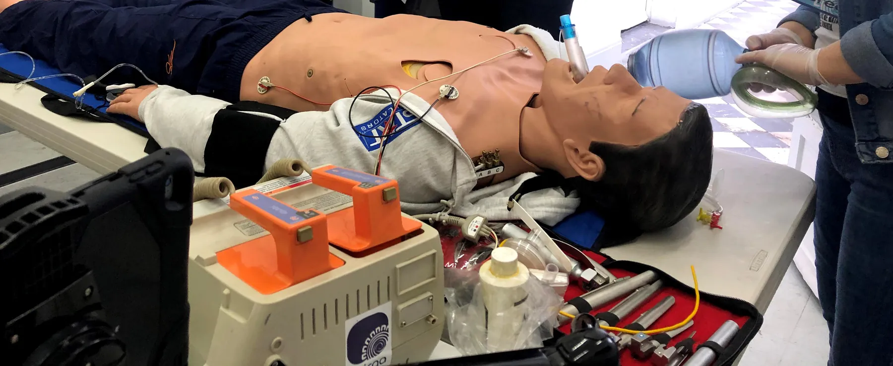 Estación de ACLS: maniquí avanzado en camilla con monitor-desfibrilador, electrodos y bolsa-válvula; solo se ven las manos de quien practica.