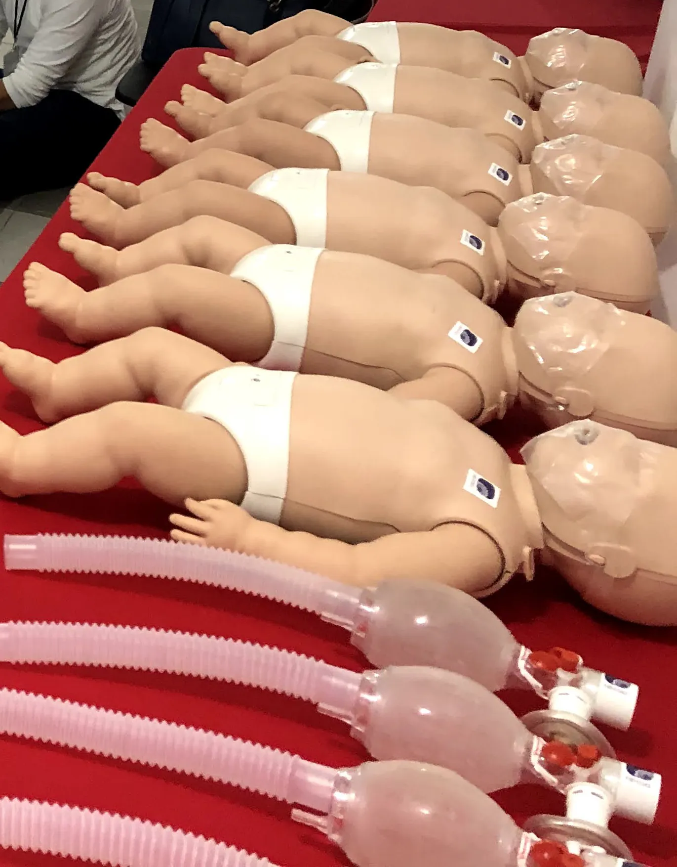 Fila de maniquíes de lactante sobre una mesa roja, cada uno con su bolsa-válvula-mascarilla pediátrica, listos para la práctica de RCP.