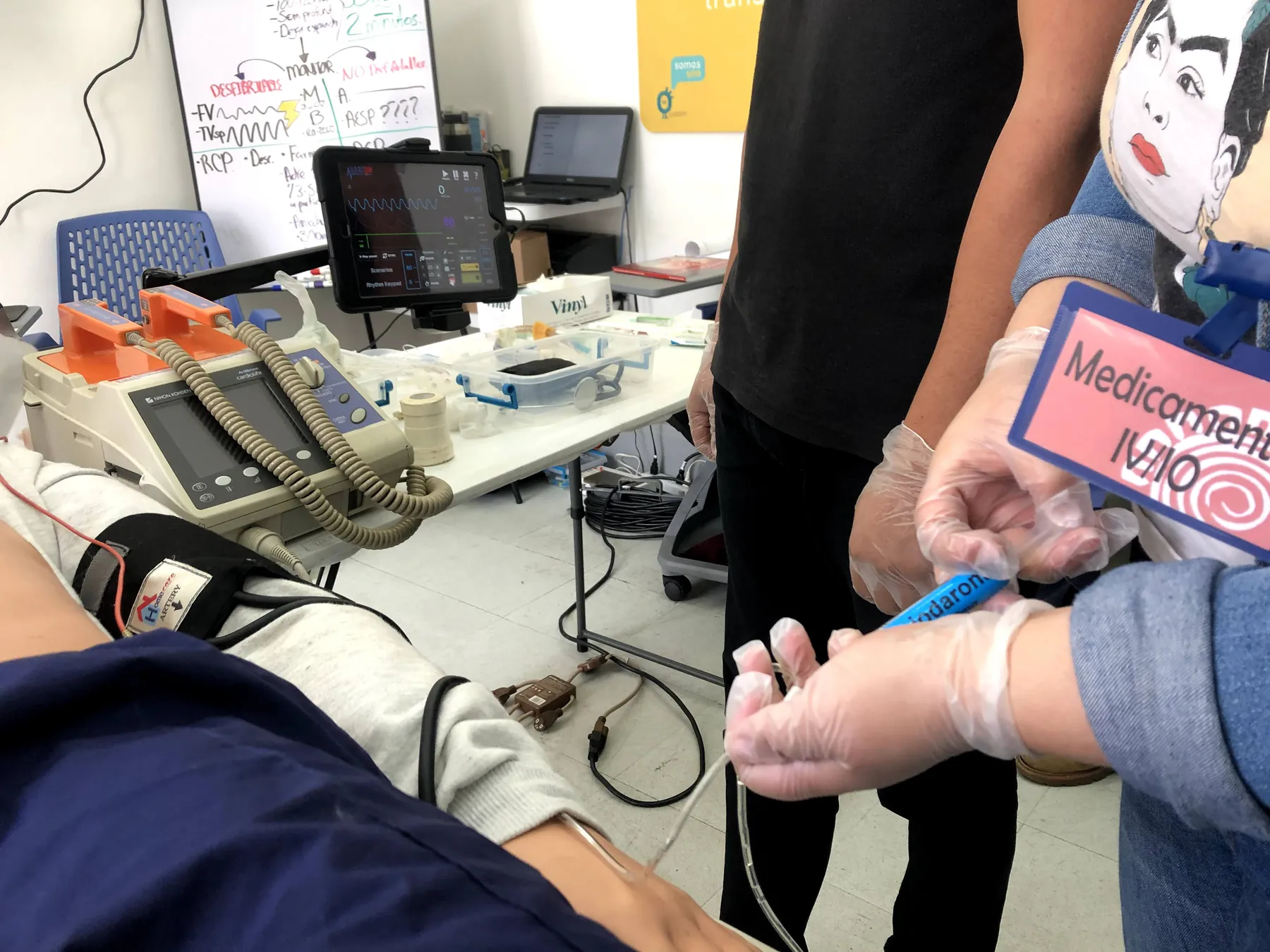 Estación de medicamentos de ACLS: manos con guantes preparan un medicamento de práctica junto al monitor-desfibrilador y al simulador de ritmos.