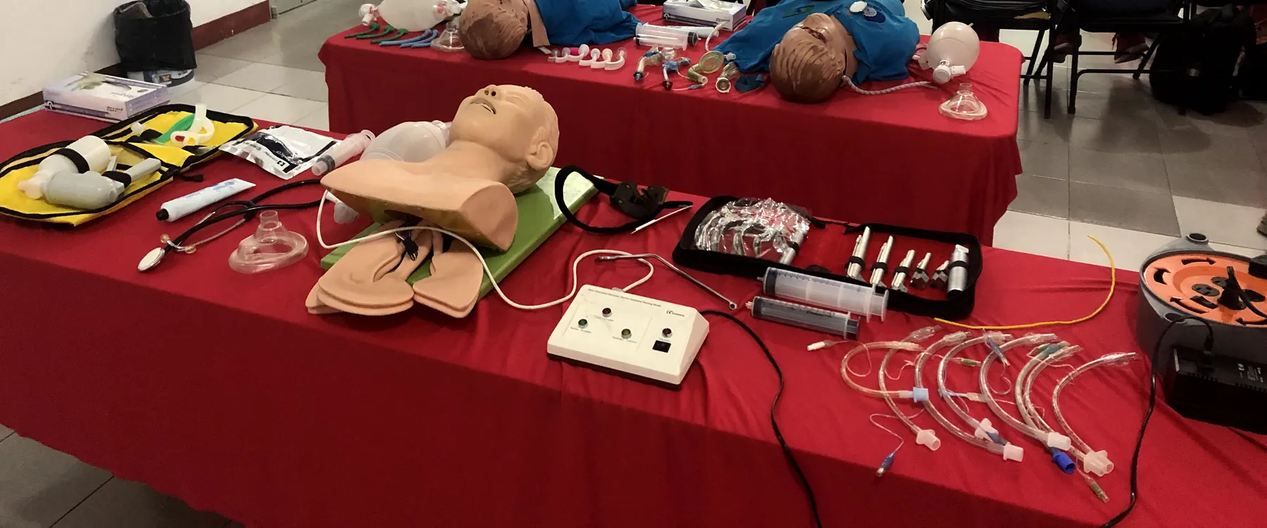 Taller de vía aérea: mesas rojas con maniquíes de intubación, laringoscopios, cánulas, tubos endotraqueales y bolsas-válvula-mascarilla.