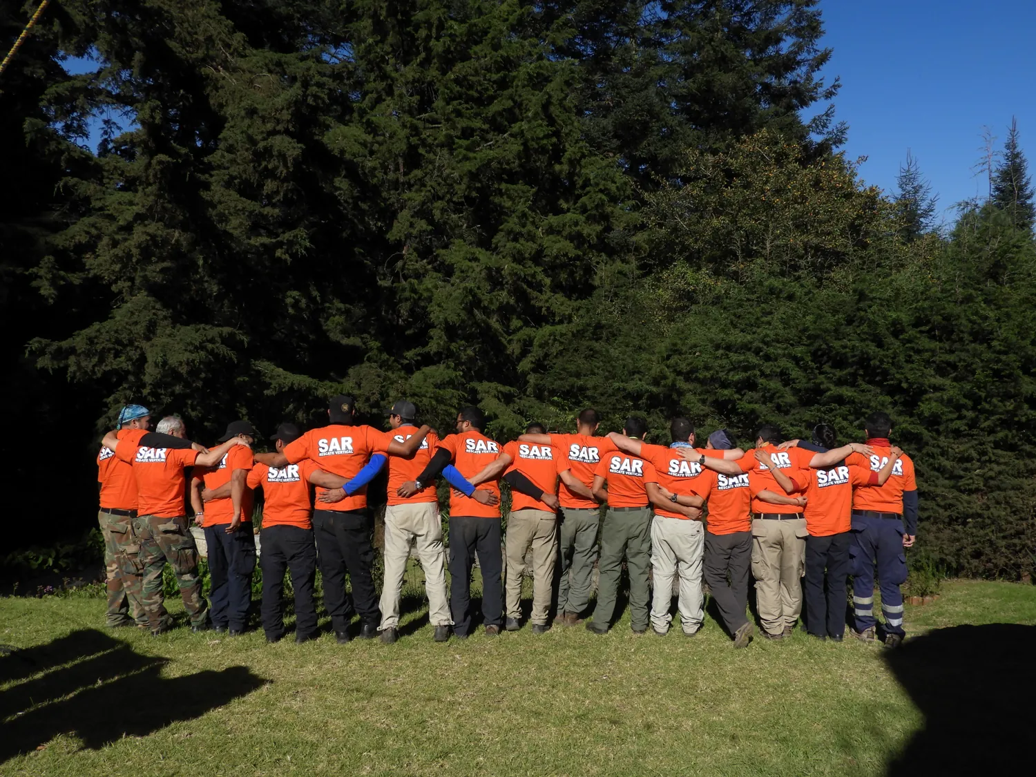 Unas veinte personas abrazadas de espaldas, con playeras naranjas «SAR · Rescate vertical», en un claro del bosque.