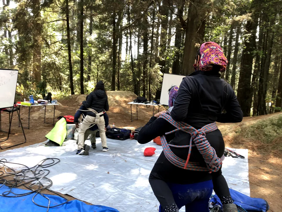 Una rescatista carga a otra persona con una silla improvisada de cuerda, de espaldas, entre pinos y rotafolios.