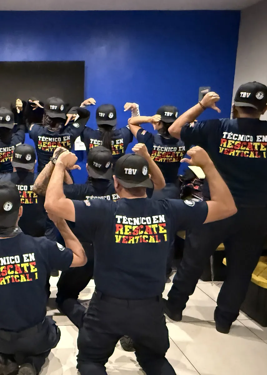 Alumnos de espaldas con playeras «Técnico en Rescate Vertical 1» y gorras «TRV», señalando su espalda frente a una pared azul.