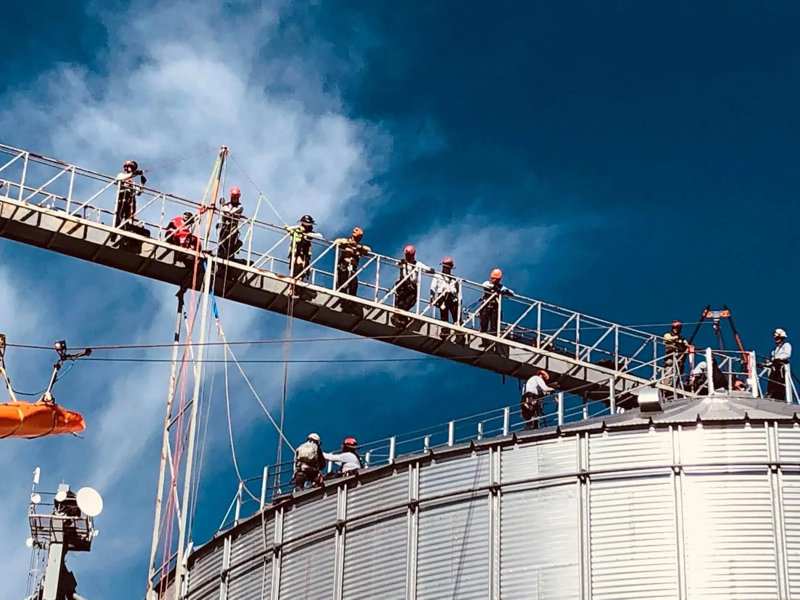 Fila de rescatistas con casco en la pasarela entre silos, contra el cielo azul; abajo cuelga una camilla naranja.