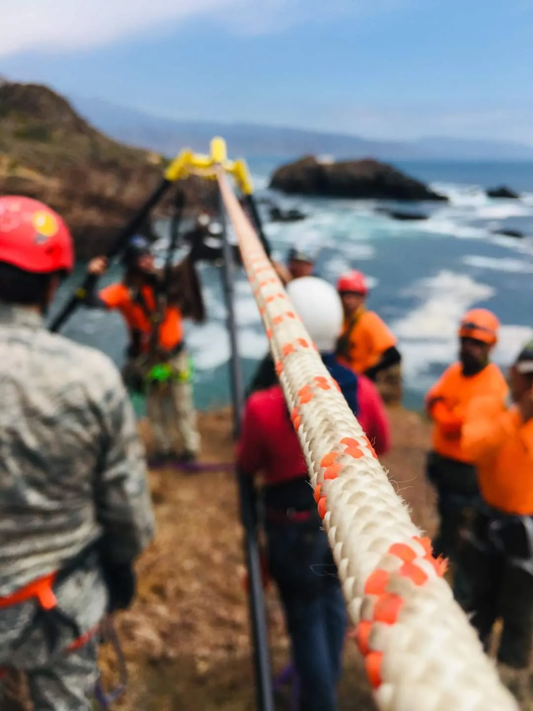 Cuerda de rescate tensada hacia el mar; al fondo, rescatistas con casco rojo y ropa naranja.
