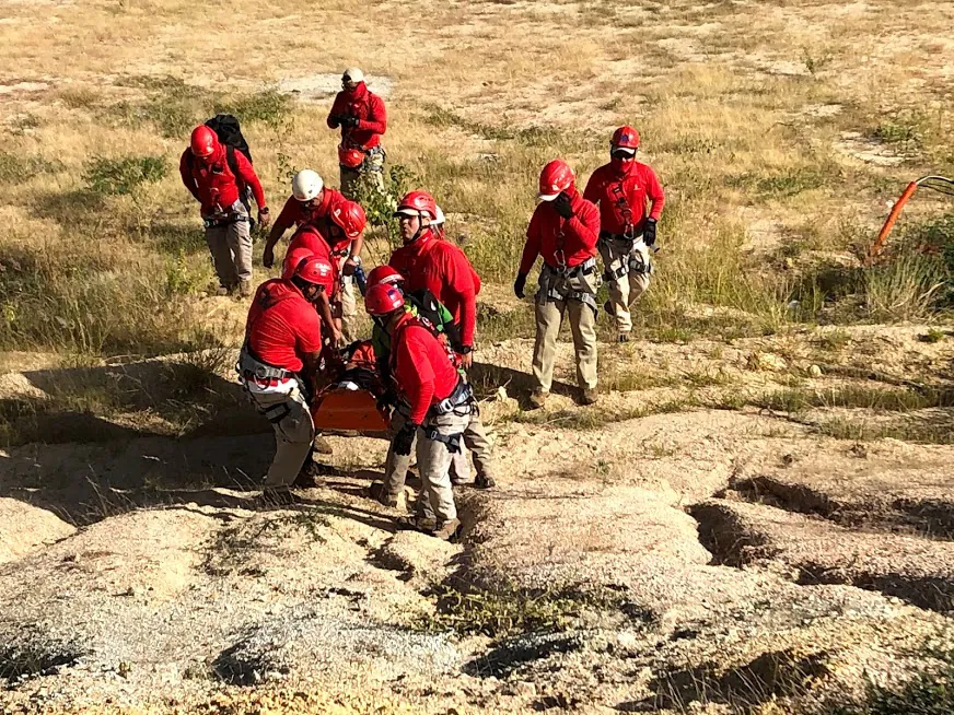 Equipo con uniforme y casco rojos transporta una camilla por terreno rocoso y árido.