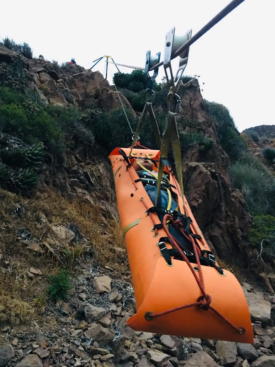 Camilla naranja tipo canastilla suspendida de una tirolesa sobre una ladera rocosa.